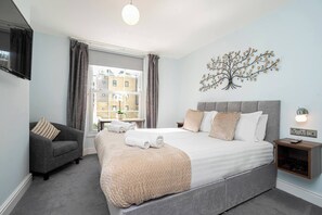 Una habitación de hotel moderna con una cama grande, un sofá gris, una ventana con cortinas, un adorno de árbol en la pared y una mesita de noche con lámpara.