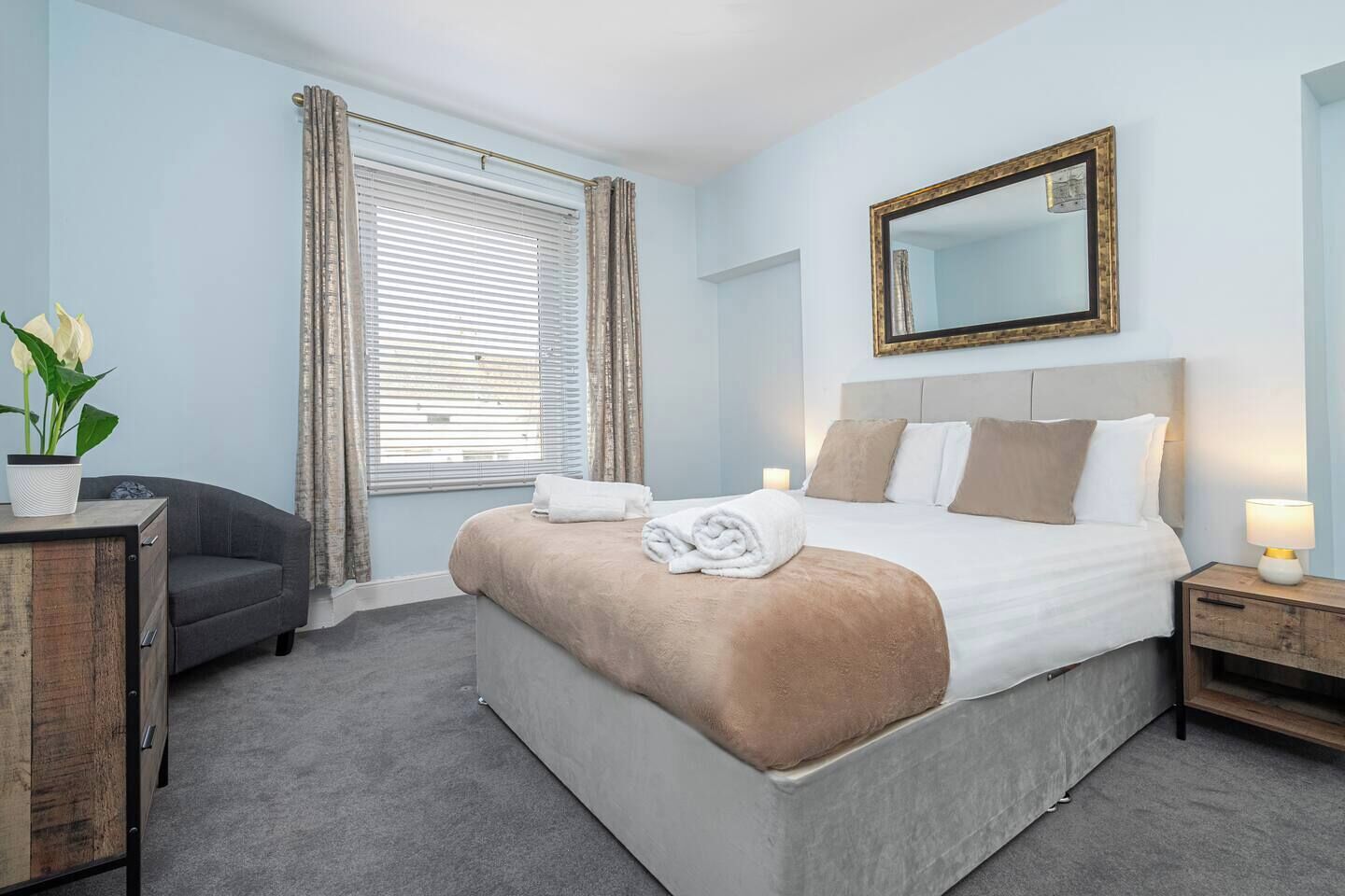 Quarto de hotel com cama, mesa de cabeceira com abajur, cômoda, janela com cortinas e um vaso de planta.