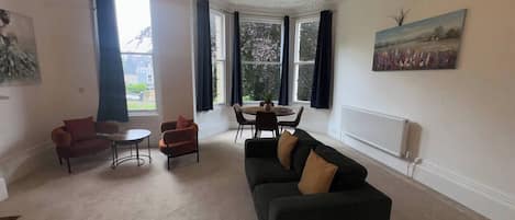 A furnished living room with a sofa, chairs, a dining table, and a view through large windows.