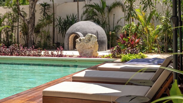 Una terraza de madera con sillones de descanso y sombrillas que da a una piscina, rodeada de plantas tropicales y una estructura de piedra.