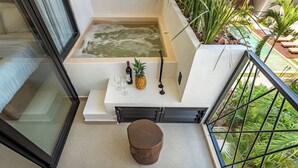 Un balcón con un jacuzzi, una mesita con una piña, vino y copas, y vistas a un jardín exuberante.