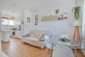 A compact living space with a kitchenette, a dining area with a glass table, a sofa, and a hanging plant. - Vacation Apartment 'Popcorn Beach House' with Shared Pool, Balcony and Wi-Fi (Corralejo)
