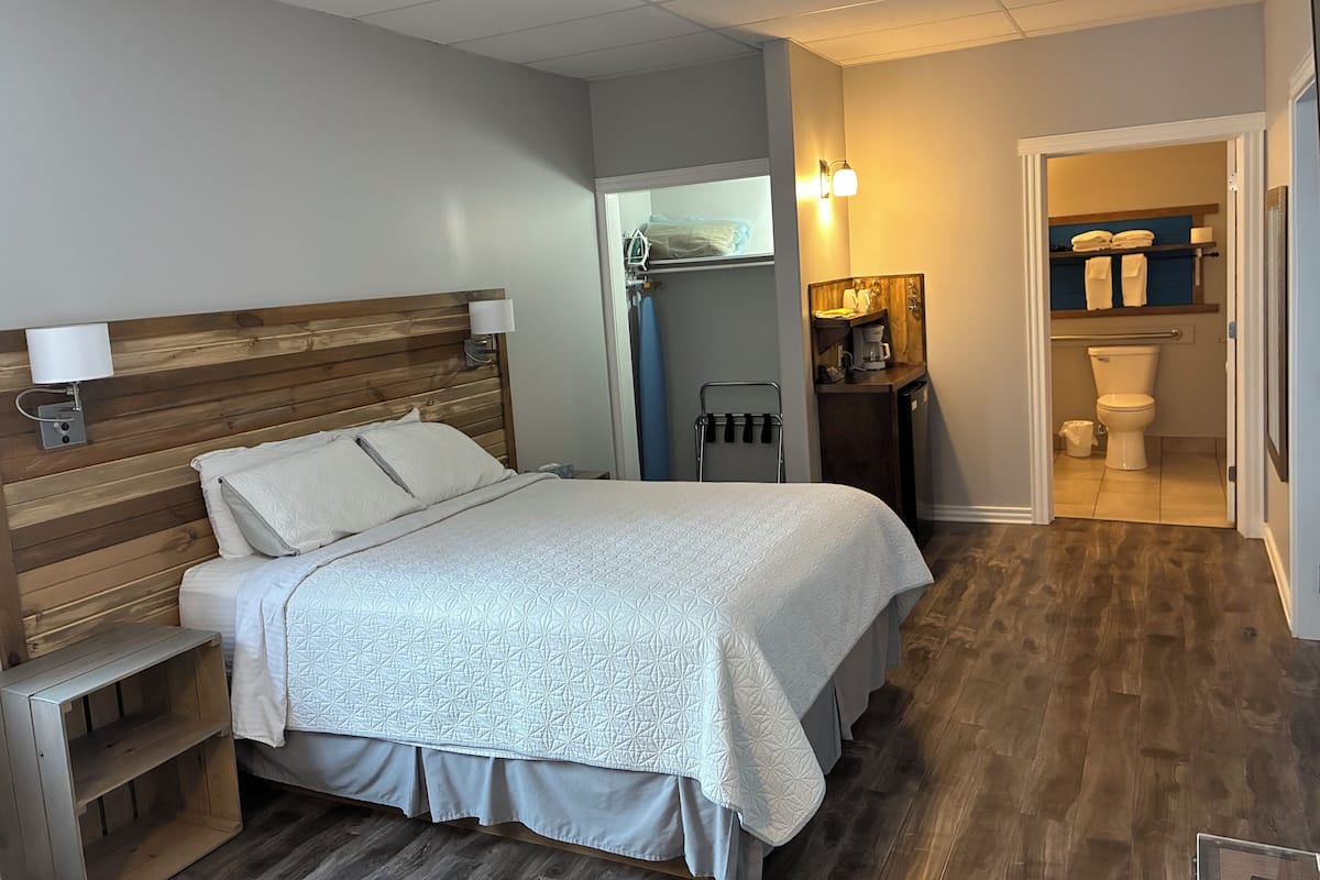 Una habitación de hotel con cabecera de madera, una cama con colcha blanca, una mesita de noche, una pequeña estantería con artículos de aseo, una cafetera y vistas al baño.