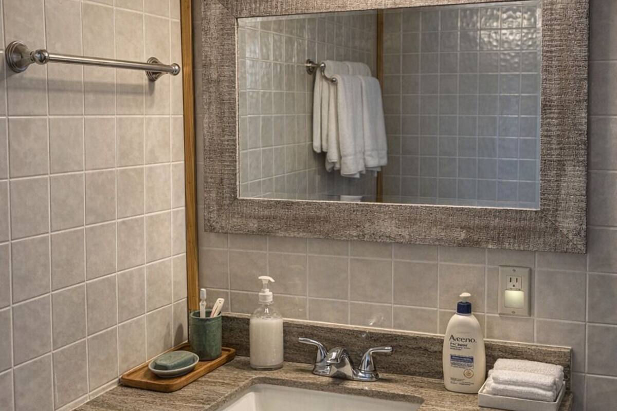 Un baño con lavamanos de encimera de granito, un espejo con marco texturizado, un perchero con toallas blancas y un dispensador de jabón.
