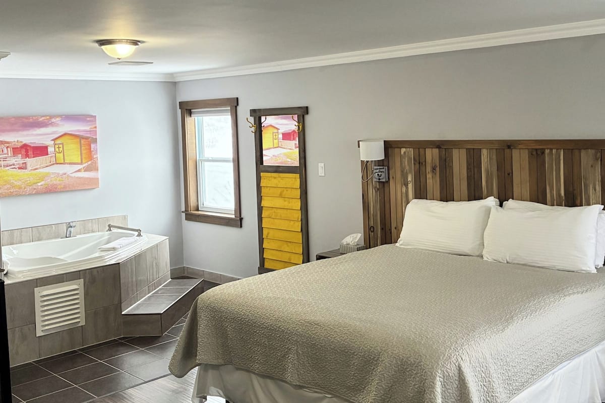 Un dormitorio con una cama grande, cabecera de madera, mesita de noche, lámpara de pared y ventana con vistas a un edificio amarillo.