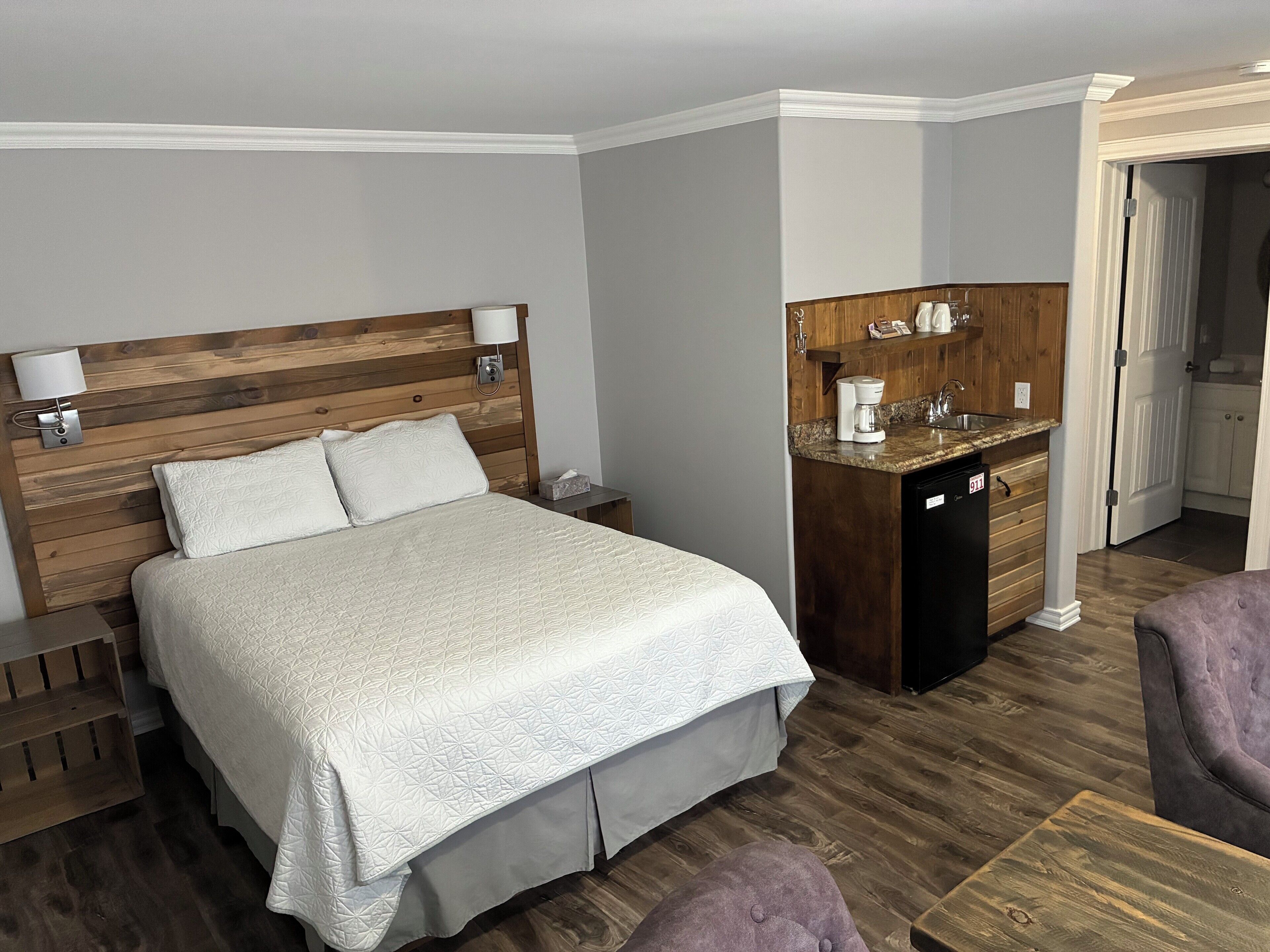 A hotel room with a wooden headboard bed, a small kitchenette, and a seating area with a purple chair.