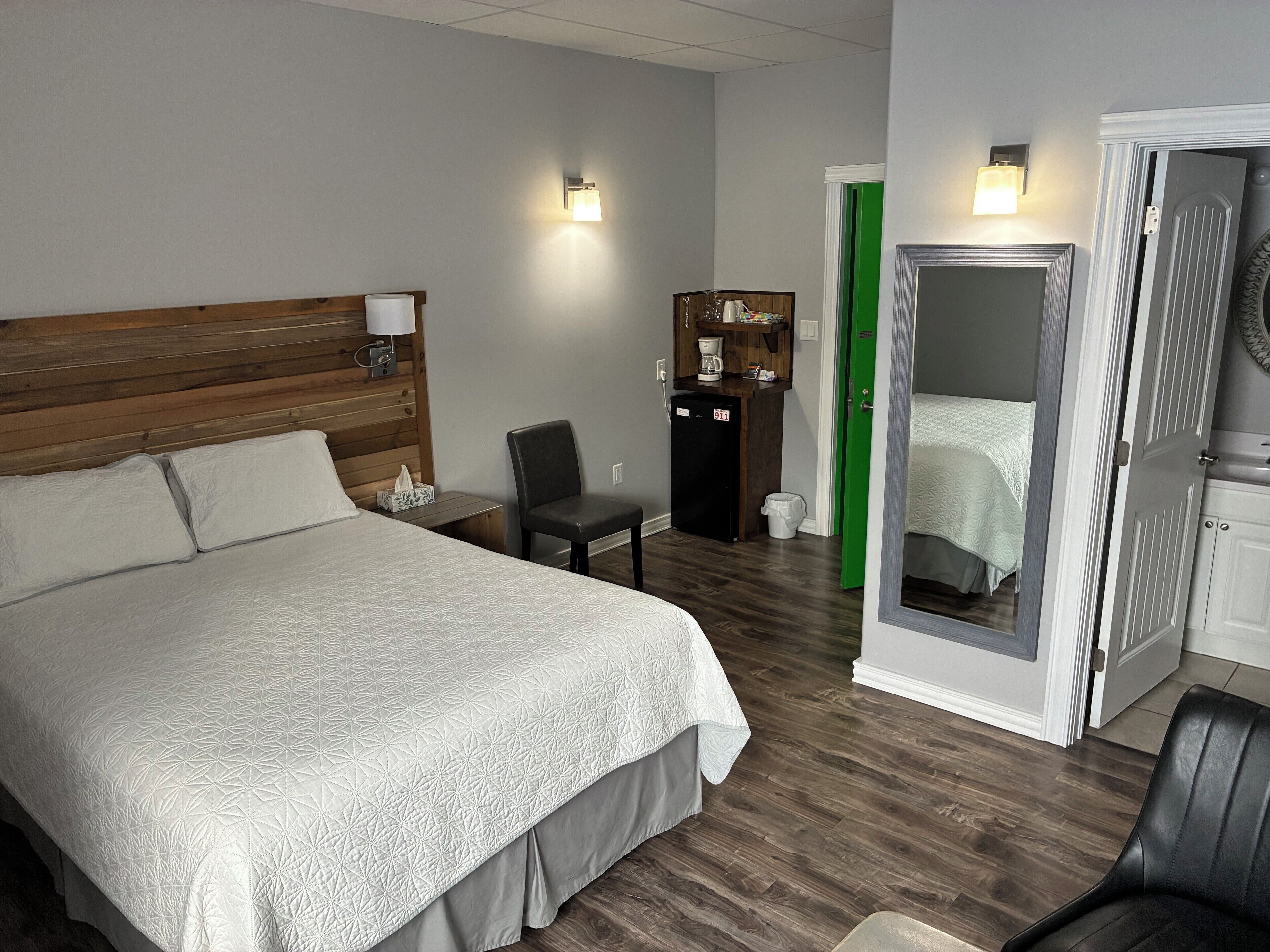 Une chambre d’hôtel avec un grand lit, une tête de lit en bois, un bureau avec une lampe, un petit réfrigérateur, une porte verte et un miroir.