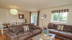 A living room with two brown sofas, a wooden coffee table, a dining area with white chairs, and a large window with curtains. - James Ville Marina Lodge 14 (Brigg)