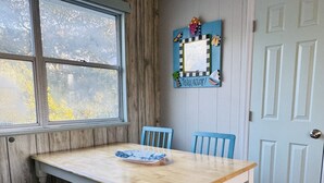 Ruokailutilassa on puinen pöytä ja siniset tuolit, ikkunasta avautuu näkymä puihin, ja ovessa on vaaleansininen paneeli.