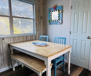 Ein Essbereich mit Holztisch und blauen Stühlen, ein Fenster mit Blick auf Bäume und eine Tür mit hellblauem Feld.