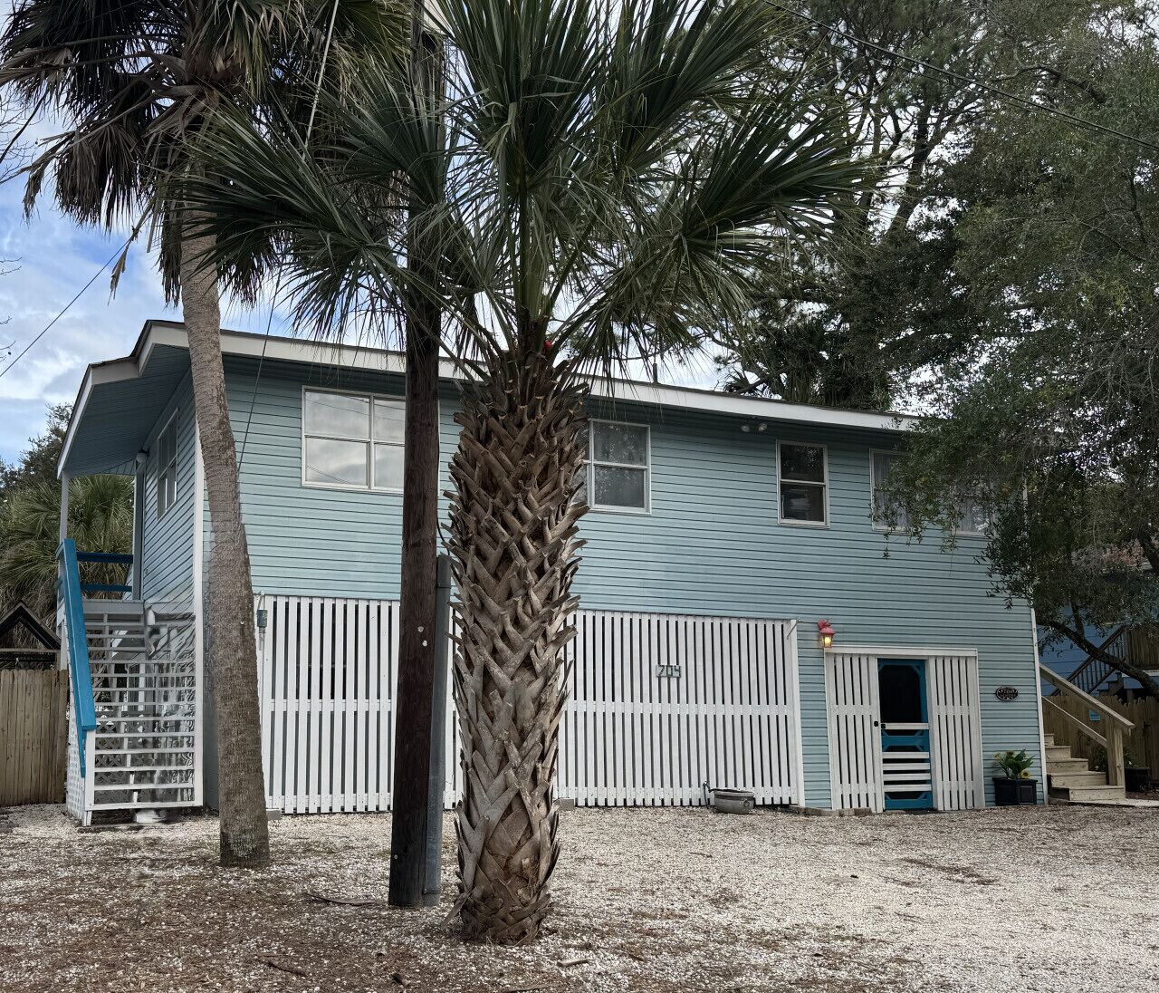 A light blue house with a white picket fence, a palm tree, and a small porch.