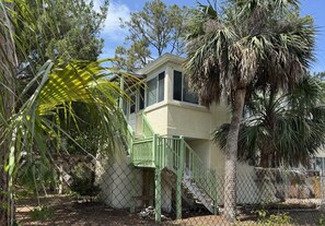 A two-story house with a green staircase leading to a balcony, surrounded by palm trees and a chain-link fence. - Sea You Soon on 12th St! One block to the beach (Tybee Island)