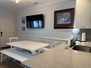 A kitchen with a white table and bench, a flat-screen TV, a framed picture, and a sink with a modern faucet. - Better Waves Ahead- 500 yds to the beach, Amenities Galore! (Tybee Island)