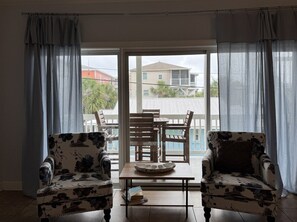Une pièce comprenant deux fauteuils, une table en bois et une vue sur un espace extérieur aménagé avec des chaises et un bâtiment, accessible par une porte-fenêtre coulissante.
