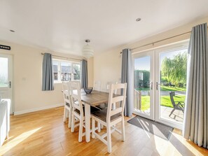 Un coin repas avec une table en bois et des chaises blanches, de grandes fenêtres et une vue sur un jardin.