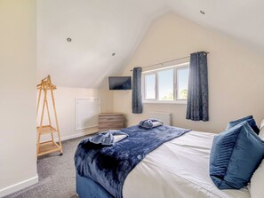 Une chambre avec un plafond en pente, un lit recouvert d’une couverture bleue, une échelle en bois, un téléviseur et une fenêtre avec des rideaux.