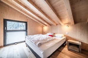 Ein Schlafzimmer in einer Holzhütte mit Bett, Nachttisch und Fenster mit Blick auf Bäume.