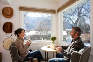 Deux personnes sont assises à une petite table ronde, avec une plante en pot, dans une pièce dotée d’une grande fenêtre et où des chapeaux sont accrochés au mur.