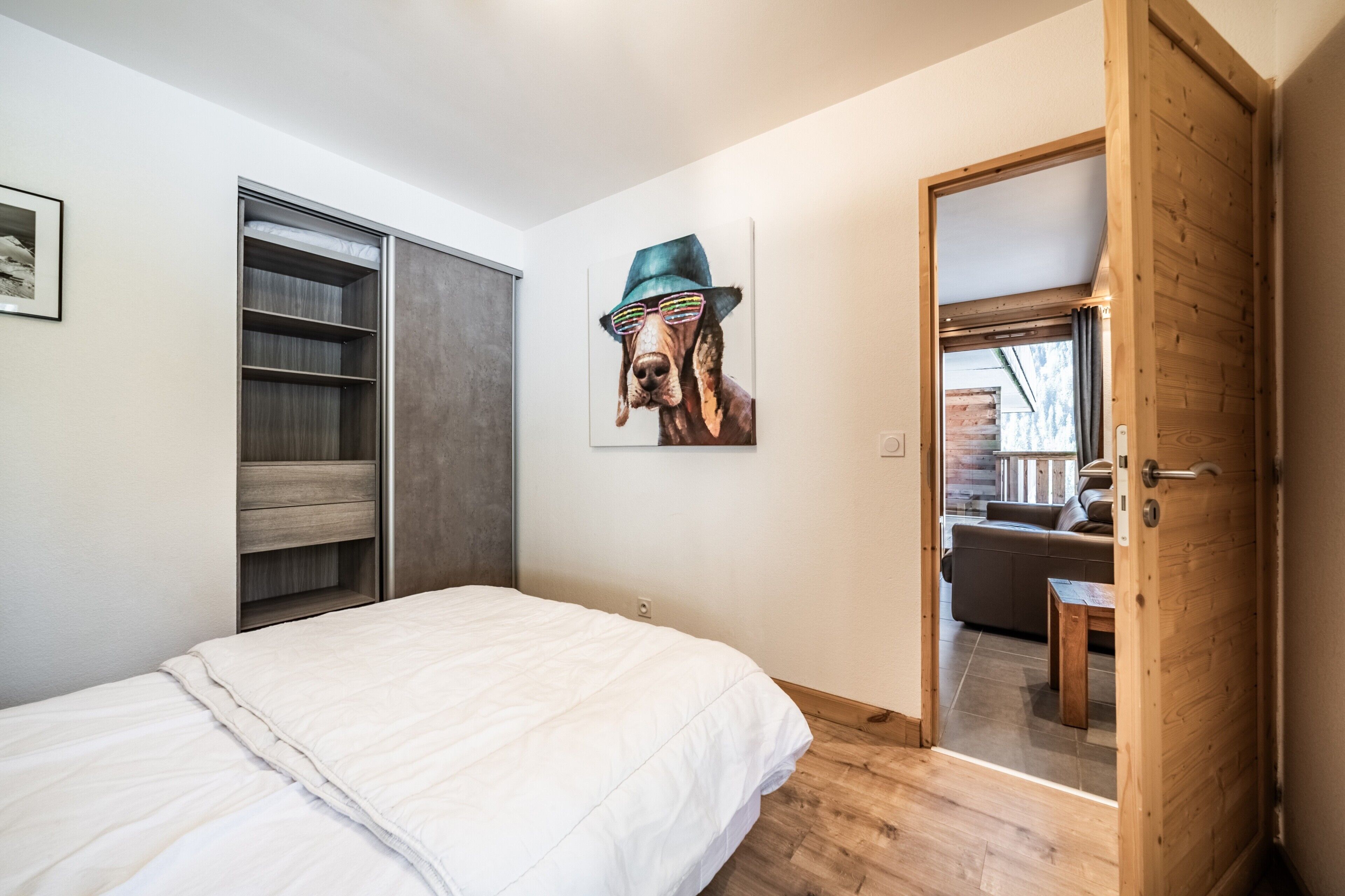Ein Schlafzimmer mit Bett, Schrank, einem Hundebild und einer Tür zu einem Wohnbereich.