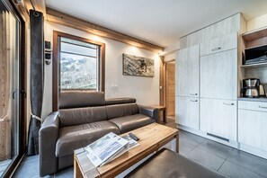 Ein modernes Wohnzimmer mit Ledersofa, Couchtisch mit Zeitschriften, einem Gemälde an der Wand und einer Küchenzeile mit Schränken und Geräten.