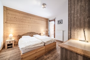 Une chambre à coucher avec une tête de lit en bois, deux lits, une table de chevet avec une lampe, un banc en bois et une fenêtre donnant sur un beau paysage.