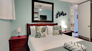 A neatly made bed with a green bedspread and tropical leaf-patterned pillows, a wooden nightstand with a red lamp, and a mirror above the bed. - Maui Parkshore 116 (Kihei)