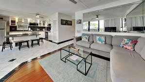 A modern living room with a grey sofa, a glass coffee table, and a kitchen area with white cabinets and a built-in oven. - Maui Parkshore 116 (Kihei)