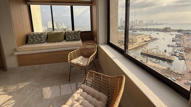 Una habitación moderna con un ventanal grande, un banco acolchado y sillones de mimbre.