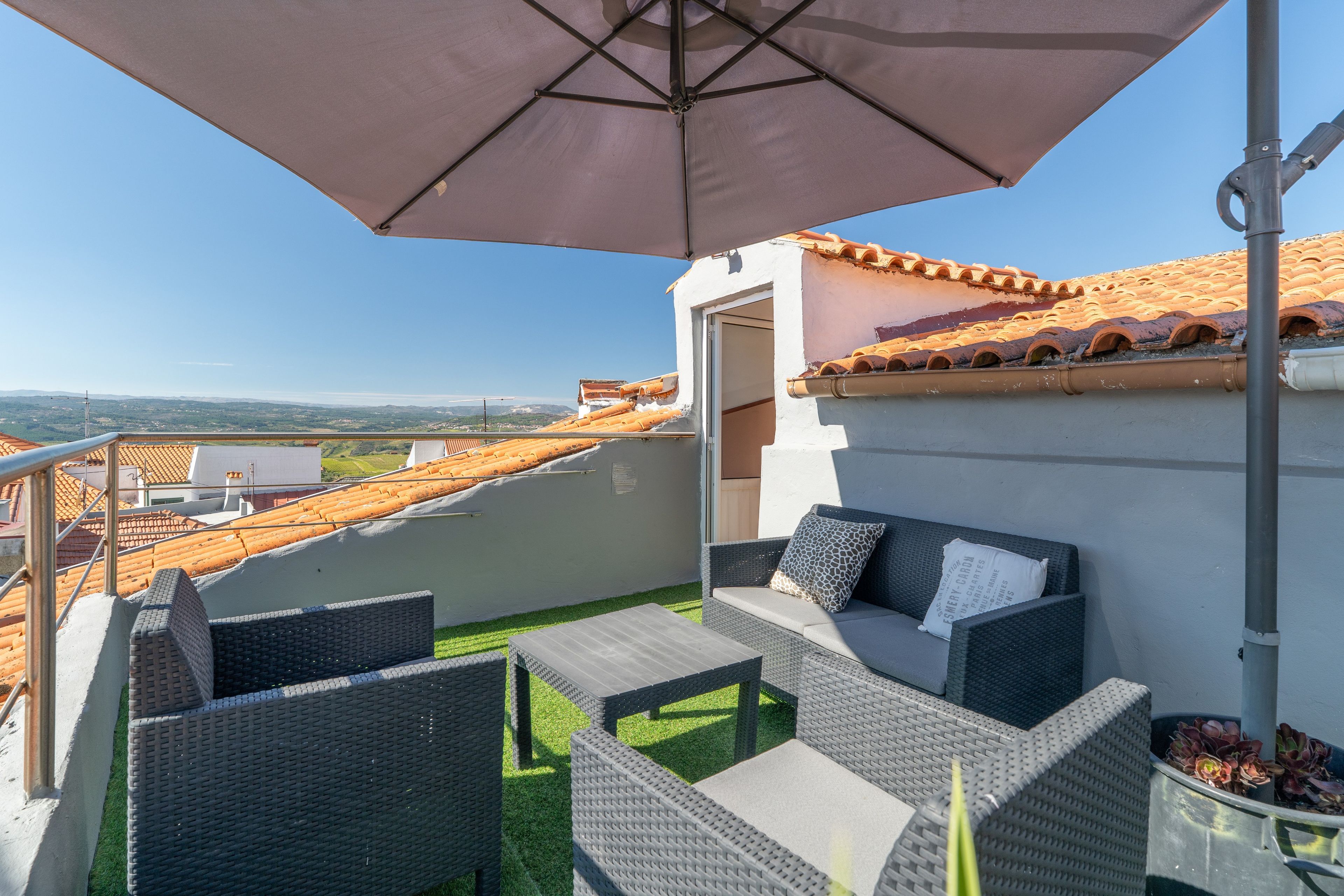 A balcony with wicker furniture, a view of terracotta rooftops, and a clear sky.