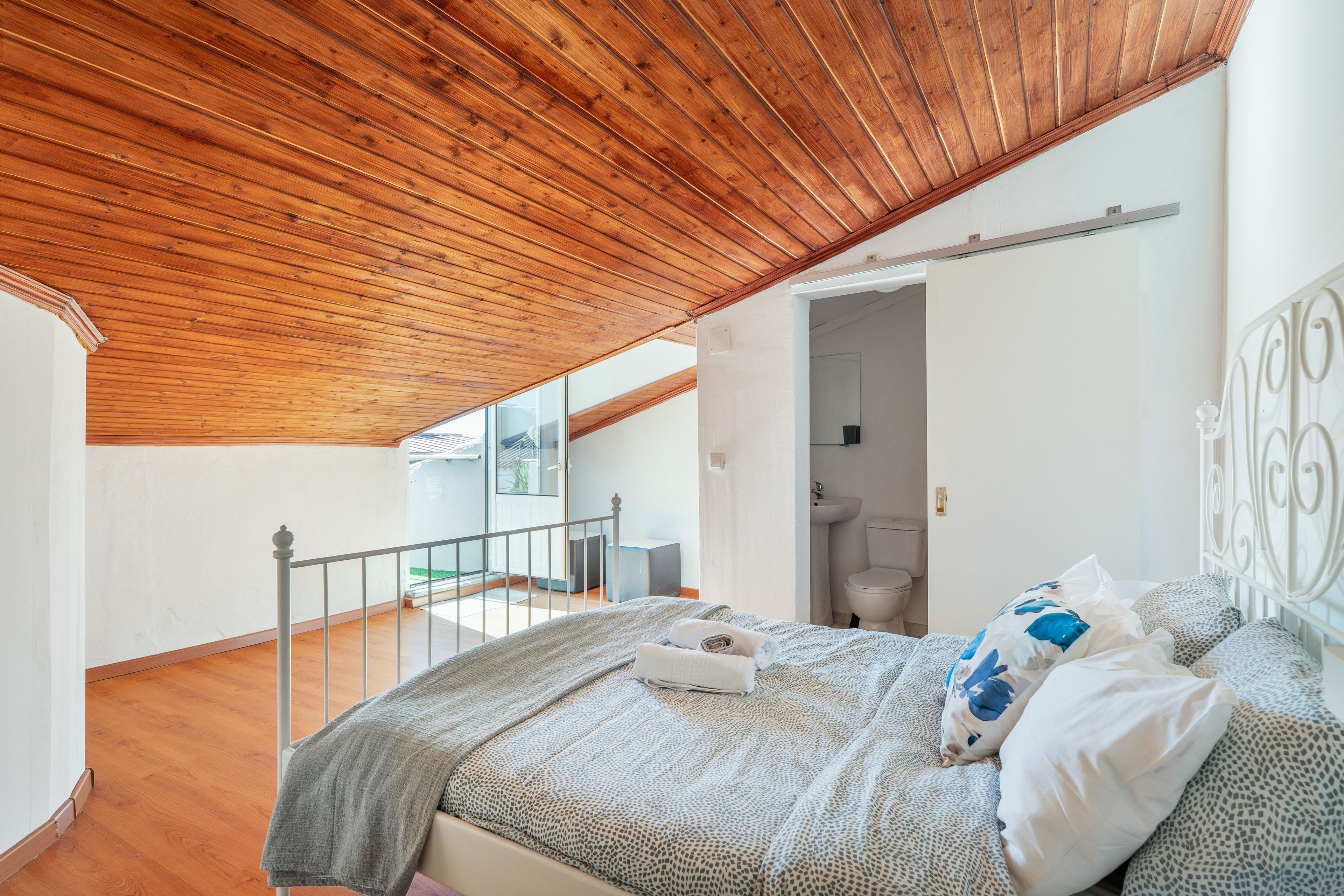 Un dormitorio con techo de madera, una cama con ropa de cama estampada, una mesita de noche blanca y vistas al exterior.