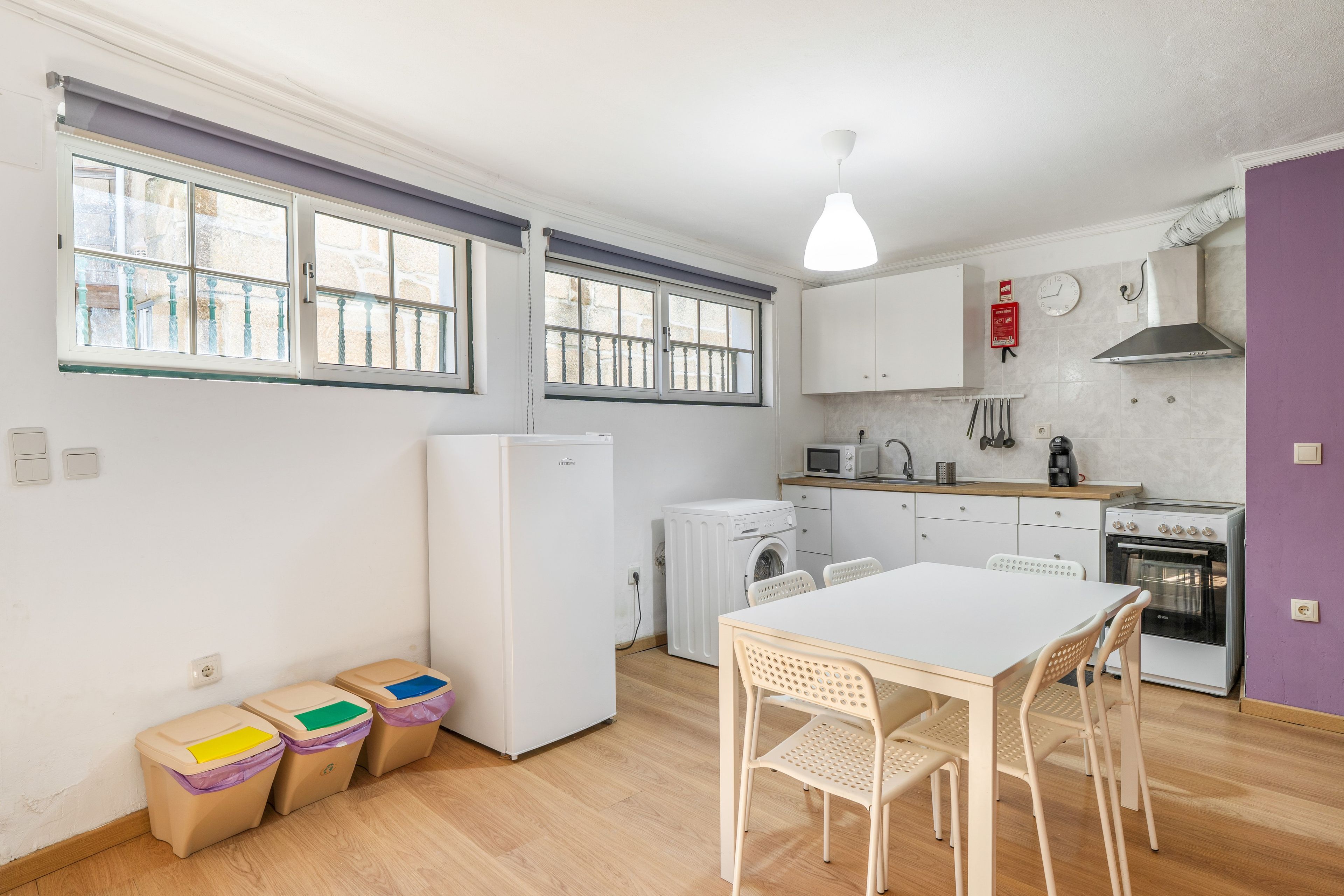 A compact kitchen with a washing machine, a microwave, and a stove. There are two windows, a table with chairs, and a trash bin.