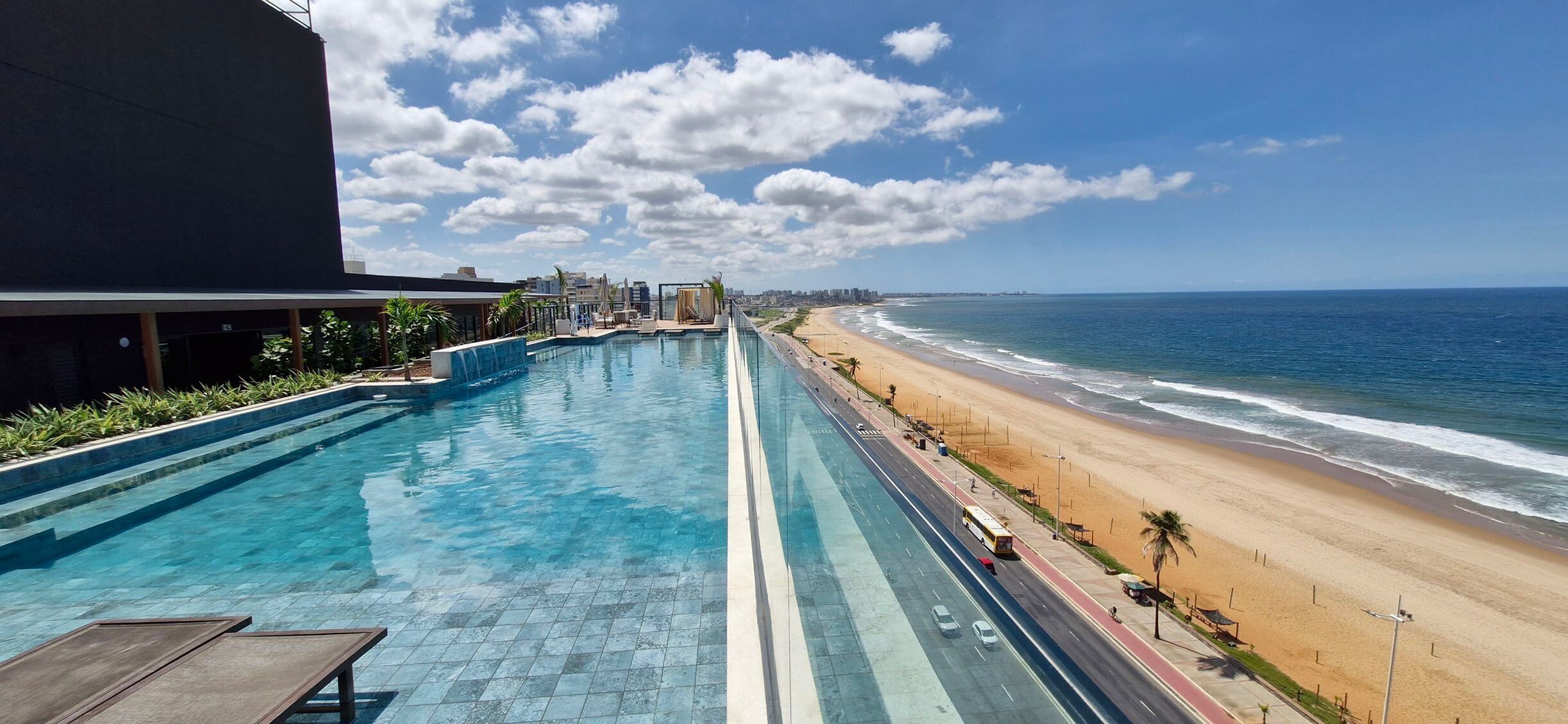 A long swimming pool adjacent to a beach with a road running parallel to the water.