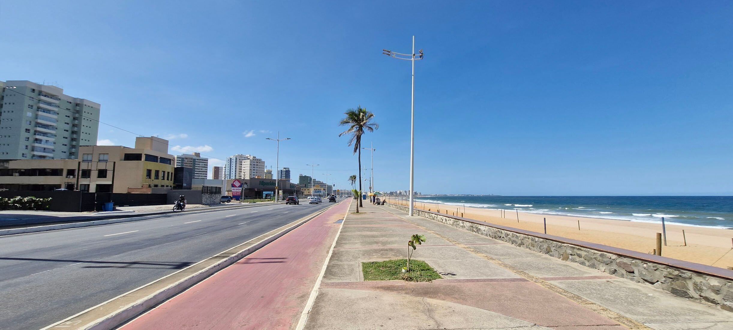 Una carretera costera con un carril bici rojo, acera y una playa al fondo.