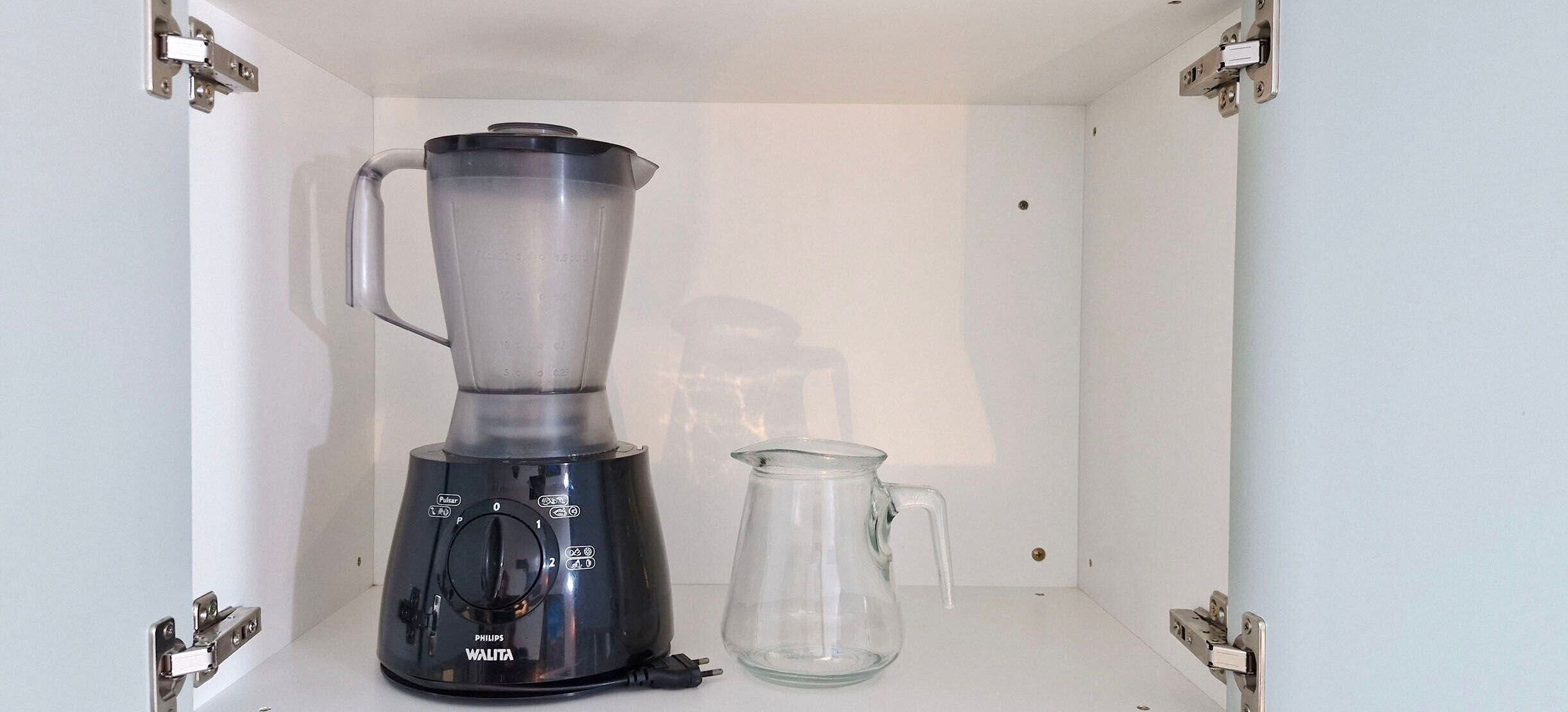 A black Philips blender with a glass pitcher, a clear glass pitcher, and a white shelf in a white cabinet.