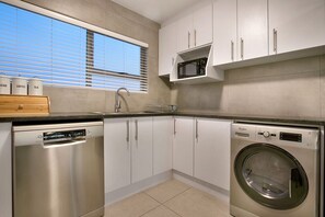 A modern kitchen with stainless steel appliances, white cabinets, and a window with blinds. - Ocean View A603 - Two Bedroom Apartment, Sleeps 4 (Cape Town)