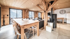 A wooden interior with a dining table, chairs, a sofa, a kitchen area, and large windows offering mountain views. - Martonne - Le Haut Apartment: Mountain View, Private Terrace & Wi-Fi (Châtel)