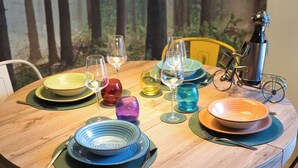 Une table ronde en bois, dressée avec différentes assiettes, bols et verres à vin de couleurs variées, accompagnée de couverts et d’une sculpture décorative représentant un vélo.