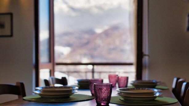 A dining table with plates and glasses, set against a window with a mountain view. - Achillea - trilocale in verde con vista montagne (Cremeno)