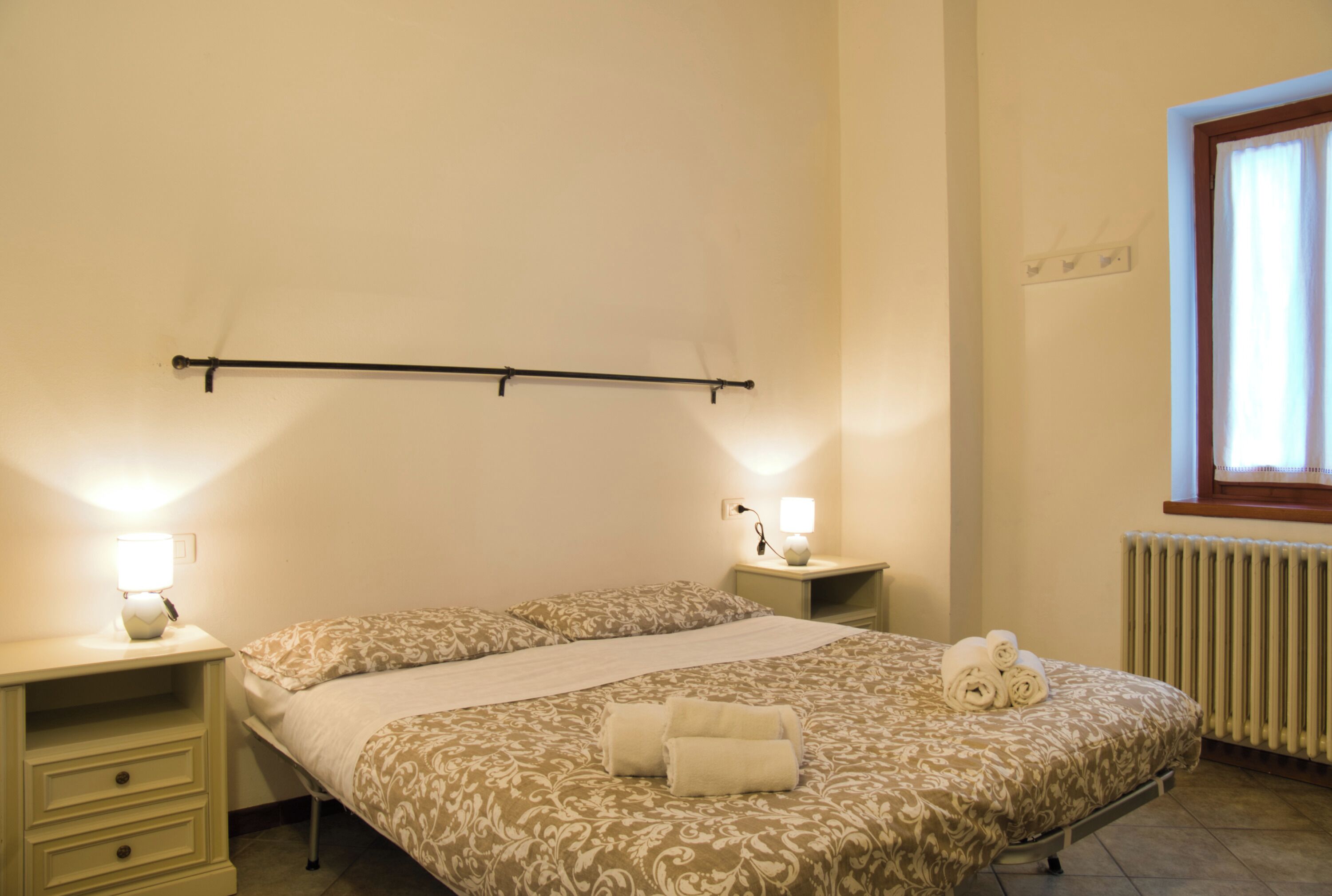 Un dormitorio con cama, dos mesitas de noche, un radiador y una ventana con cortinas.
