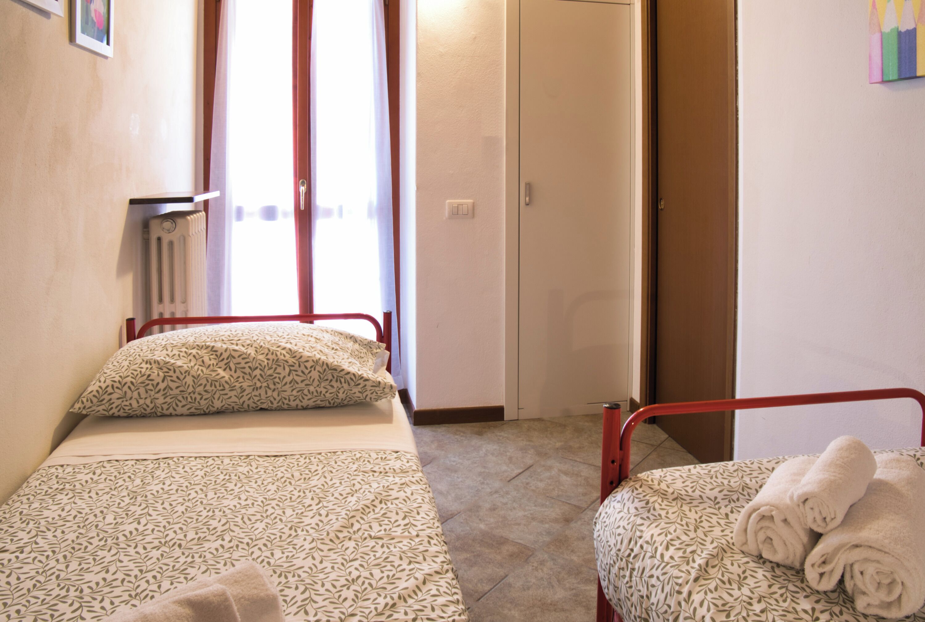 Une petite pièce bien éclairée, avec deux lits simples, chacun agrémenté de draps et d’oreillers à motifs. On y trouve un radiateur au mur, une porte donnant sur une autre pièce et un luminaire fixé au mur.