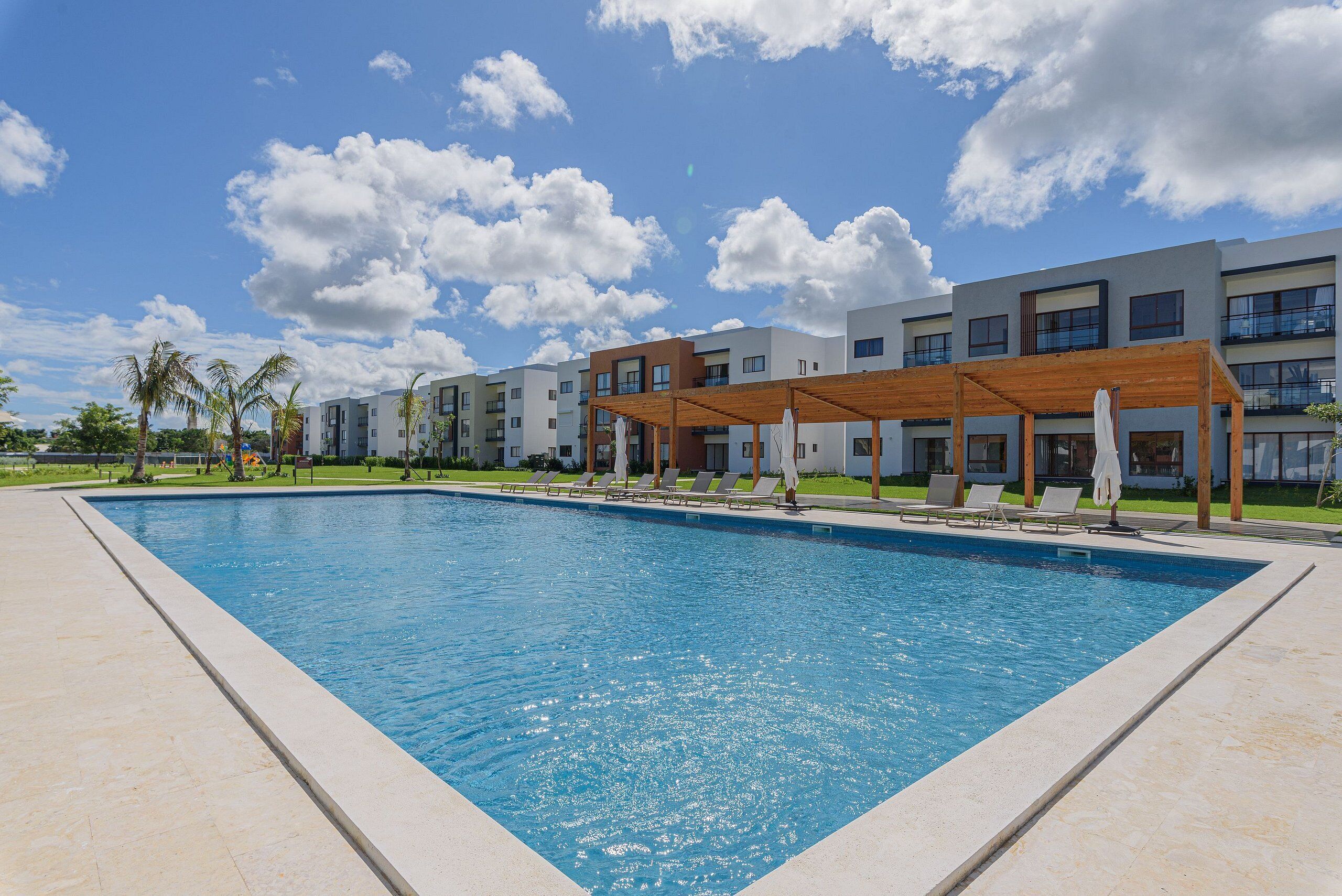 A rectangular swimming pool with a wooden pergola and lounge chairs, surrounded by modern buildings and palm trees.