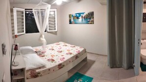 Une petite chambre avec un lit simple, dotée d’une moustiquaire, d’une fenêtre avec des volets, d’un tableau accroché au mur et d’une porte donnant sur la salle de bain.