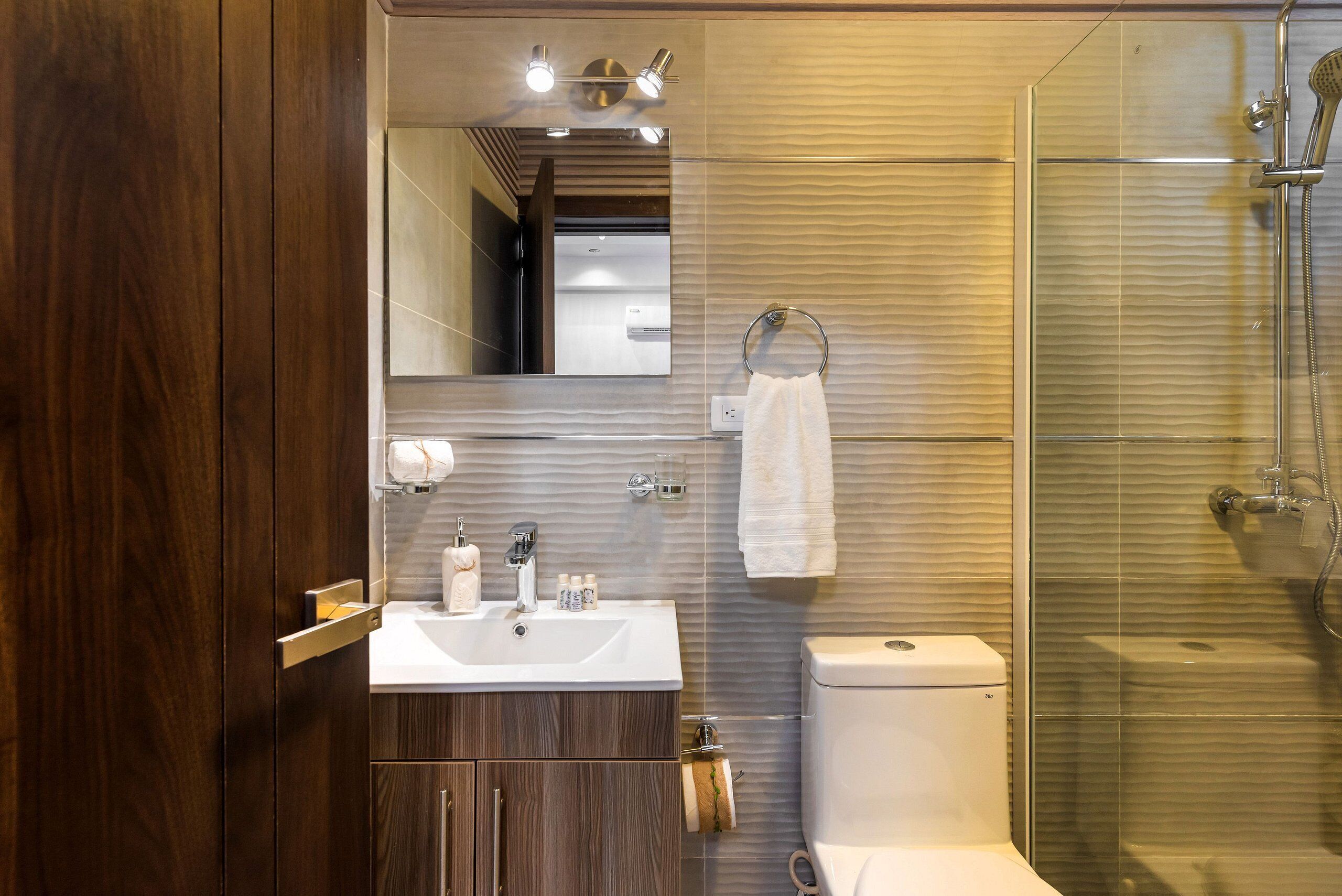 Un baño moderno con tocador de madera, un lavabo blanco, un espejo, un perchero para toallas y una ducha con mampara de vidrio.