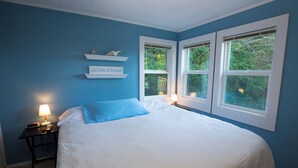 Un dormitorio con una cama grande, dos mesitas de noche con lámparas, una estantería con objetos decorativos y una ventana con vistas a árboles.