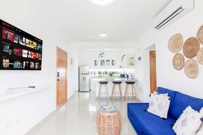 A modern living room with a blue sofa, a mounted TV displaying a Netflix interface, and a kitchen in the background. - 2 bedrooms, pool, 7 minutes from the airport (Punta Cana)