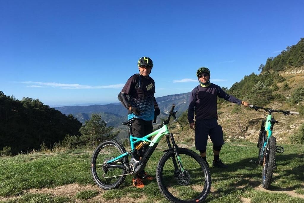 Dos personas en una colina cubierta de hierba, con bicicletas, observando un paisaje montañoso.