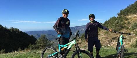 Dos personas en una colina cubierta de hierba, con bicicletas, observando un paisaje montañoso.