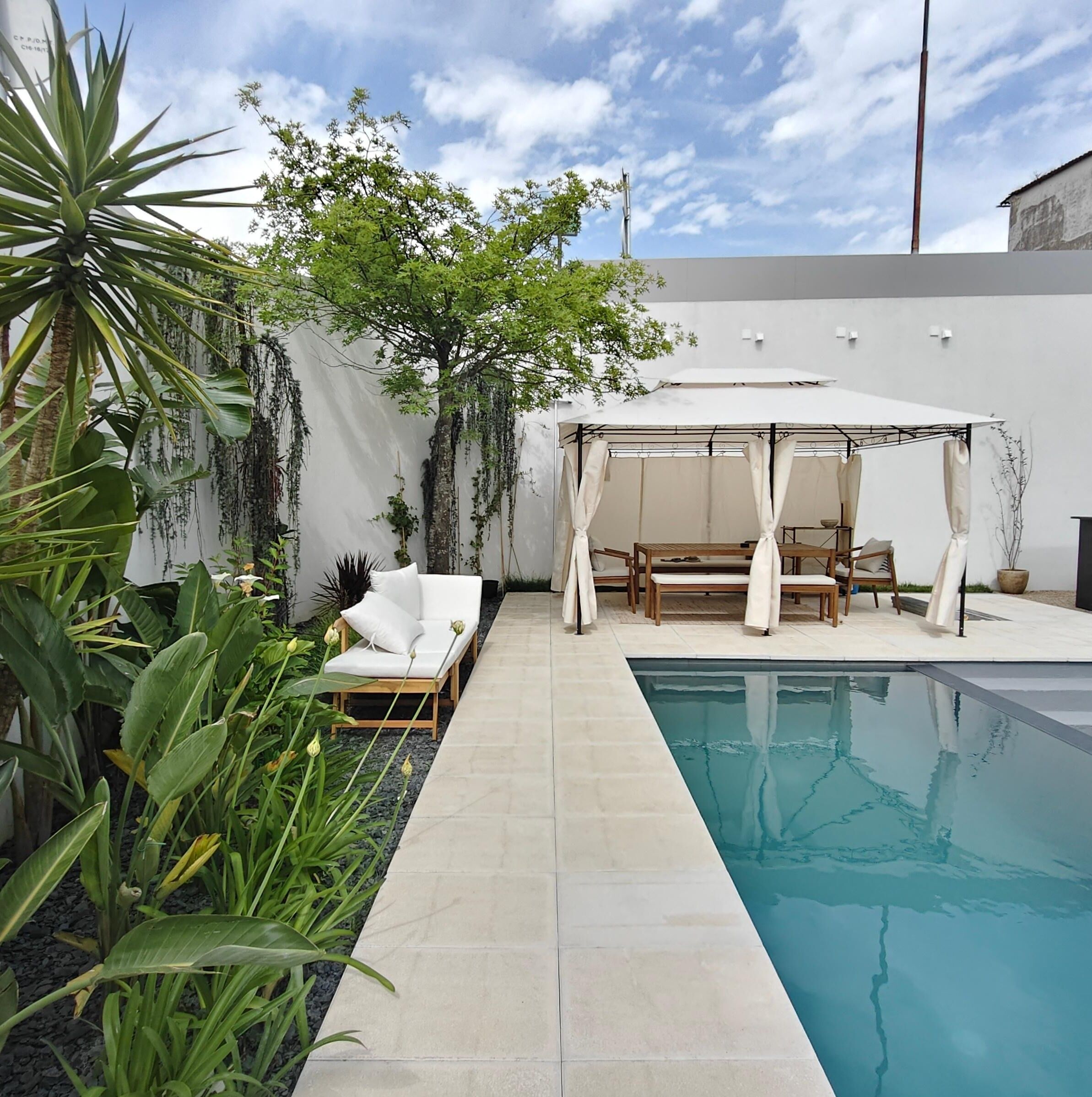 A private pool area with a white outdoor seating area, including a sofa and chairs, surrounded by lush greenery and a white wall.