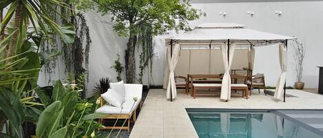 A private pool area with a white outdoor seating area, including a sofa and chairs, surrounded by lush greenery and a white wall.