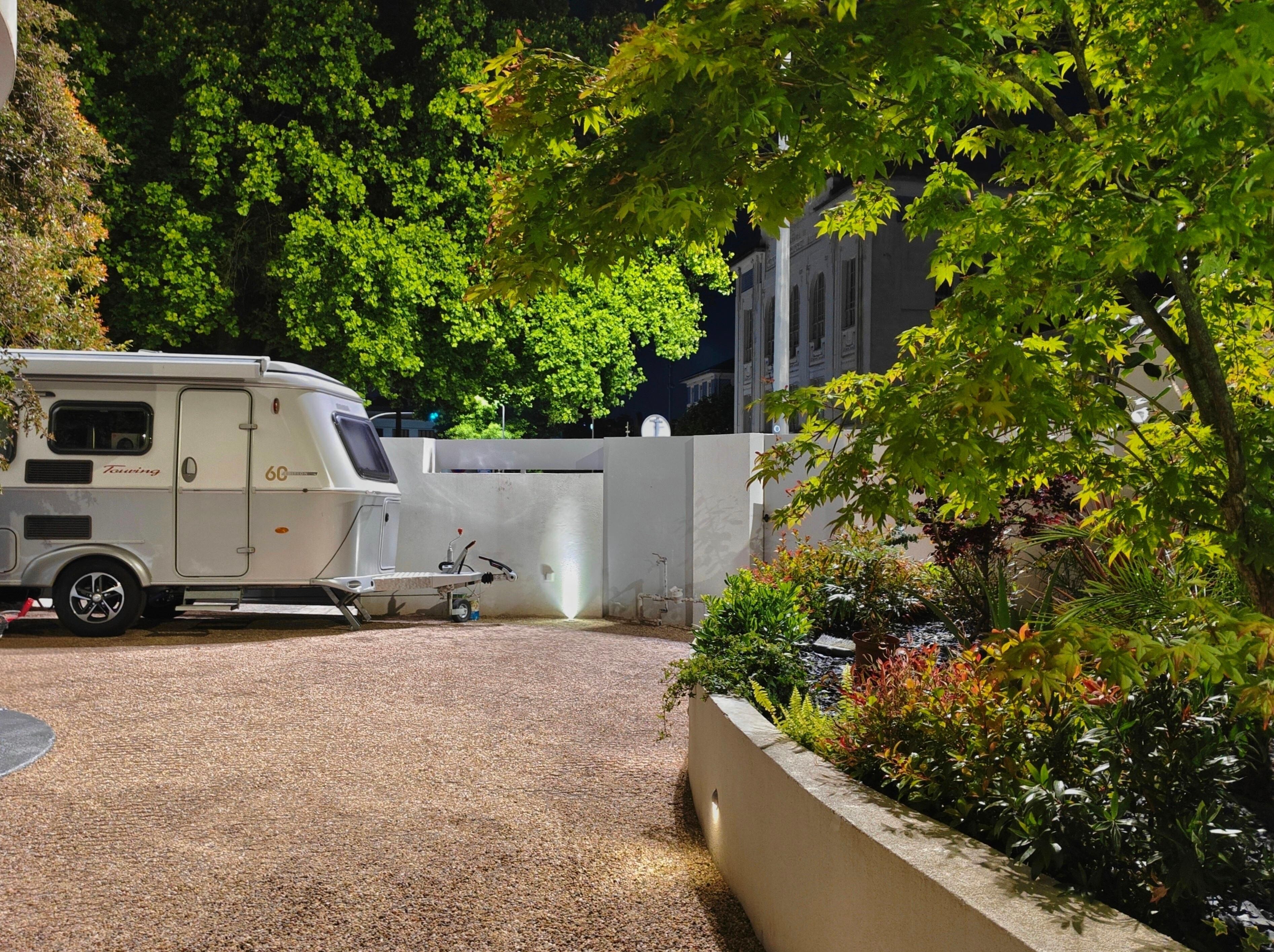 Une caravane est garée sur une allée gravillonnée, avec un jardin et un bâtiment en arrière-plan.